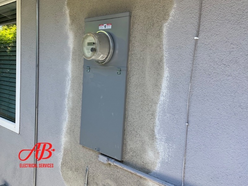 AB Electrical Services An electrical meter box is mounted on a gray wall next to a window. The initials "AB" and "Electrical Services" are visible at the bottom left of the image. Orange County CA