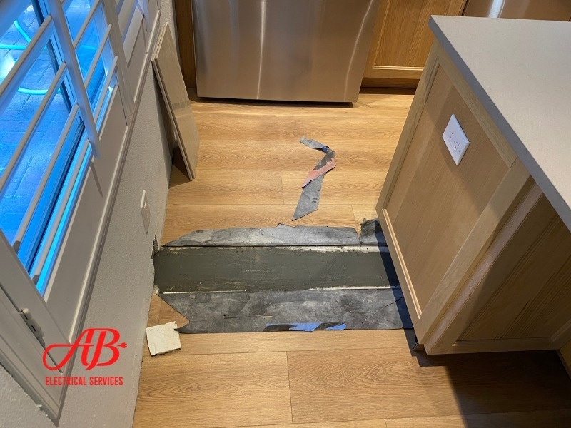 AB Electrical Services Kitchen with damaged wooden floor. Exposed subflooring visible, surrounded by intact surface. Light wood cabinets and stainless steel fridge in the background. Orange County CA