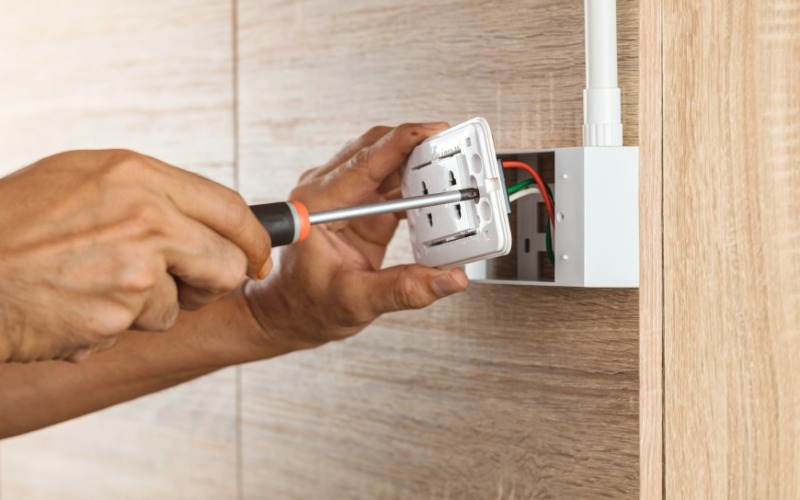 AB Electrical Services During storm season, a person uses a screwdriver to install or repair a wall electrical outlet with visible wiring, ensuring proper electronics surge protection. Orange County CA