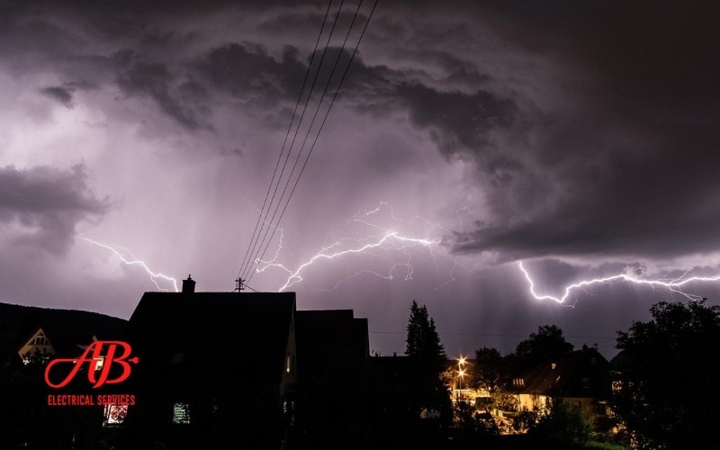 AB Electrical Services Dramatic lightning illuminates a dark, stormy sky over silhouetted houses, with electrical wires visible in the foreground—reminding homeowners of the importance of whole-house surge protection during storm season. Orange County CA
