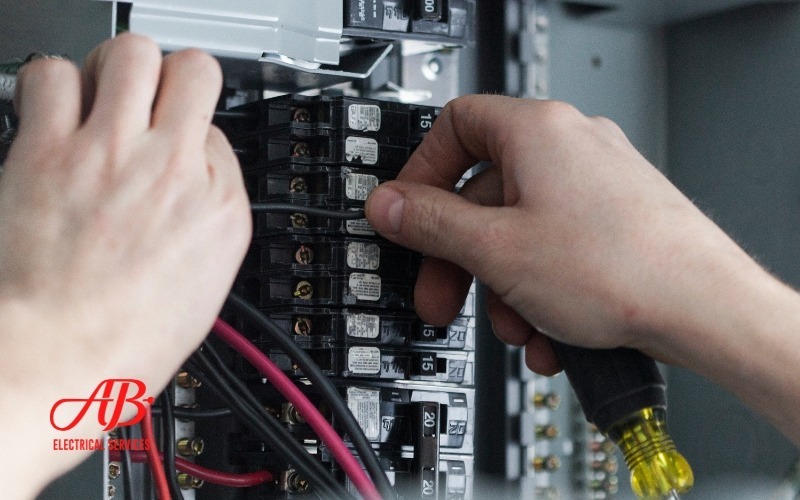 AB Electrical Services Hands skillfully adjusting wires in an electrical system panel with a screwdriver. The process ensures a safer, smarter setup for optimal performance. Logo in the corner reads "AB Electrical Services. Orange County CA