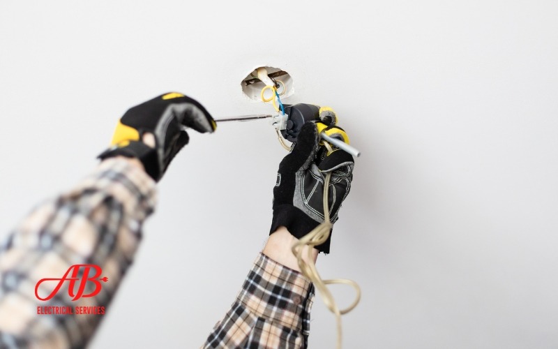 AB Electrical Services An electrician with gloved hands carefully installs wiring in a ceiling, ensuring a safer electrical system. A logo reads "AB Electrical Services" in the corner, highlighting their reliable workmanship. Orange County CA