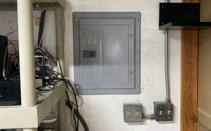 AB Electrical Services Electrical panel mounted on a wall next to shelves filled with cables and electronics; two light switches are below the panel. Orange County CA