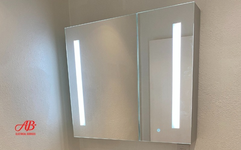 AB Electrical Services A large rectangular mirror with LED light strips on both sides is mounted on a beige wall. A logo reading "AB Electrical Services" is in the bottom left corner. Orange County CA