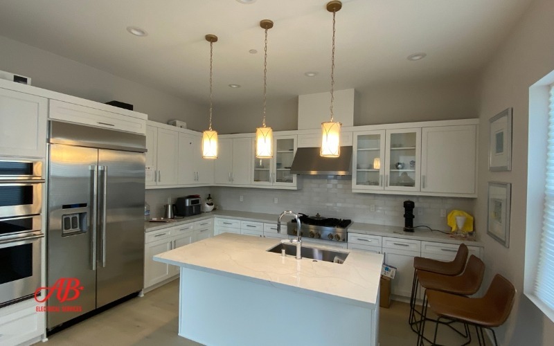 AB Electrical Services Modern kitchen with white cabinetry, stainless steel appliances, an island with a white countertop, three pendant lights, and two brown chairs. Orange County CA