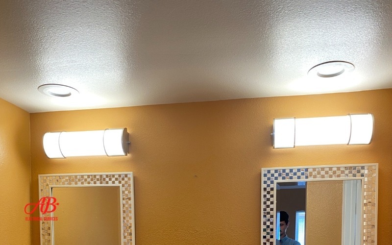AB Electrical Services Bathroom with yellow walls, two mirrors, and ceiling lights. A person is partially visible in the reflection. Orange County CA