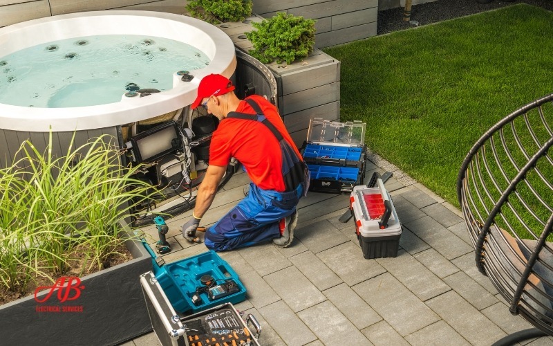 AB Electrical Services A technician in a red cap and blue overalls is meticulously working on a hot tub with toolboxes and equipment on the patio, ensuring the ultimate relaxation space. Orange County CA