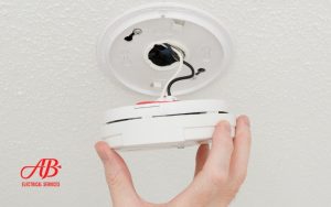AB Electrical Services A person holds a smoke detector while installing or removing it from a ceiling mount, with visible wiring and a company logo in the bottom left corner, ensuring home protection against electrical fires. Orange County CA