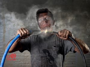 AB Electrical Services A man with a dirty face and shirt holds two sparking electrical wires, creating visible smoke and a burst of light between them—highlighting the dangers of DIY electrical repairs. Orange County CA