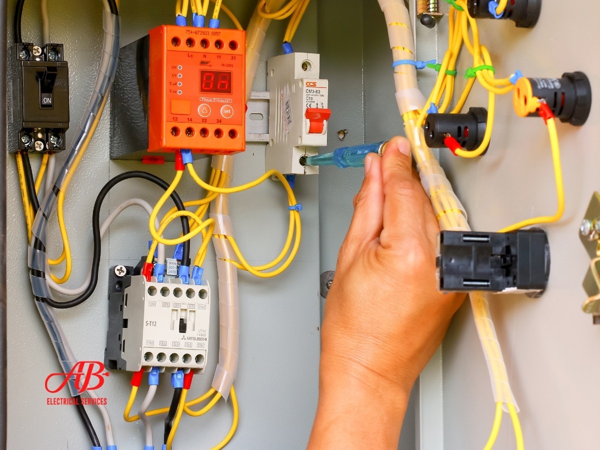 AB Electrical Services A person uses a screwdriver to adjust wiring inside an electrical control panel with various relays, wires, and an automatic transfer switch. Orange County CA