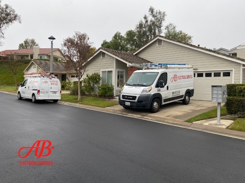 AB Electrical Services Two AB Electrical Services vans are parked outside a suburban house on a cloudy day, with tools visible on the vans’ roofs. The company logo is visible in the image corner. Orange County CA