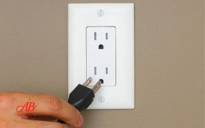 AB Electrical Services A hand is about to plug a black electrical cord into a white wall outlet mounted on a beige wall, highlighting the need for electrical outlet troubleshooting when facing outlet problems. Orange County CA