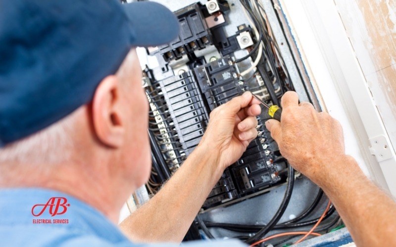 AB Electrical Services An electrician in a blue shirt and cap works on a circuit breaker panel using a screwdriver, addressing breaker box troubleshooting. The AB Electrical Services logo is visible in the corner. Orange County CA